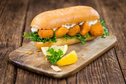 Crispy fish sandwich with lettuce and tartar sauce on a bun, served with lemon wedges on a wooden board.